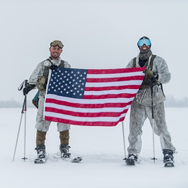 Two individuals in winter gear holding an American flag, standing in a snowy landscape.
