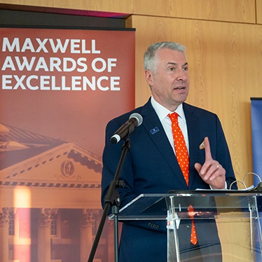 Person speaking at a podium with a "Maxwell Awards of Excellence" banner in the background.