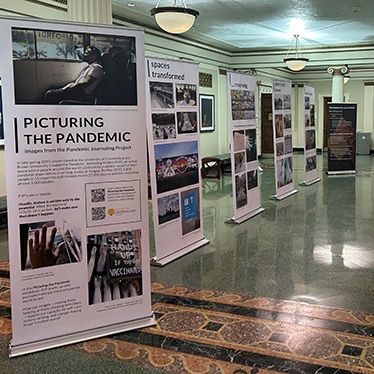 Exhibition titled 'Picturing the Pandemic' with informational panels displayed in a hallway, featuring photographs and text about the global impact of the pandemic.