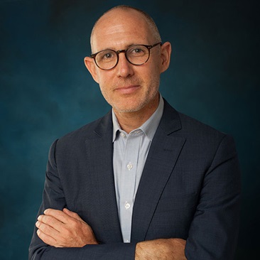 Portrait of a person wearing glasses, a dark blazer, and a light shirt, with arms crossed and standing against a dark blue background.